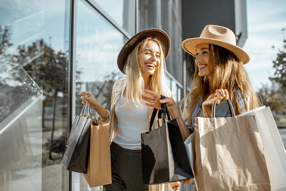 Two women wearing hats and holding shopping bags while walking and smiling outside a store with glass windows, just after discovering the best loyalty program app for small businesses.