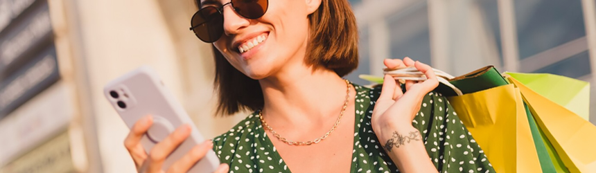 Smiling woman wearing sunglasses holds shopping bags while looking at her phone, enjoying the Best Loyalty Program App for Small Business.