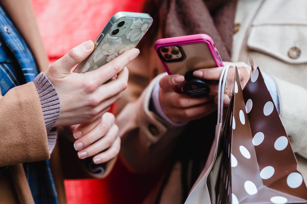 Two people holding smartphones with decorative cases; one has a floral design, the other is pink. Shopping bags are visible, perhaps indicating they've just used the best loyalty program app for small business to earn rewards.