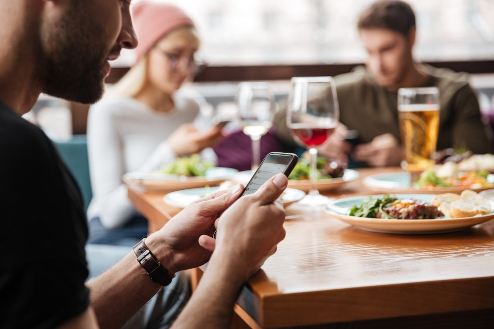 Three people sitting at a table with food and drinks, all looking at their smartphones, discussing the Best Loyalty Program App for Small Business.