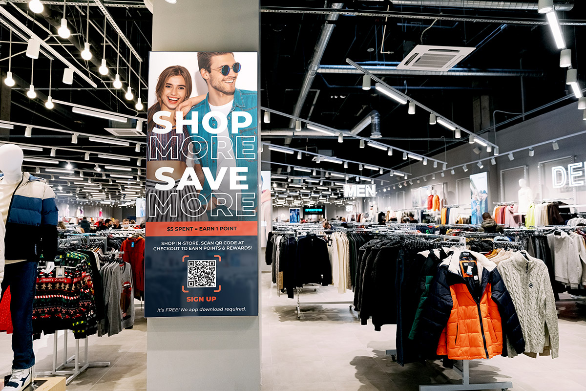 A clothing store interior with a "Shop More, Save More" promotional sign featuring a QR code and RewardPocket System Integration.