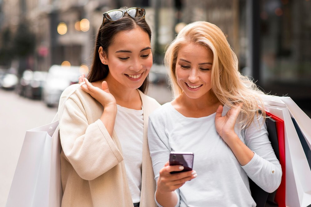 Two women with shopping bags are smiling and looking at a smartphone on a city street, excitedly discussing their latest finds and the benefits of their loyalty program.