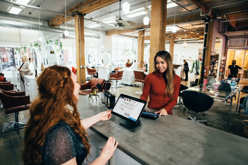 Two women smiling and conversing at a checkout counter in a stylish, open-concept salon, discussing the benefits of the new loyalty program.