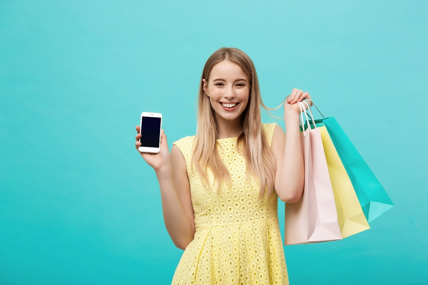 Smiling woman in a yellow dress holding shopping bags in one hand and a smartphone displaying her loyalty card in the other against a teal background.